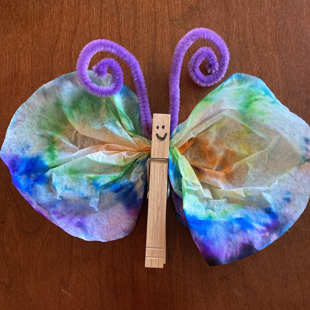 A tie-dye butterfly made from a rainbow coffee filter, a clothes pin, and a purple pipe cleaner folded in half and curled to resemble antenna.